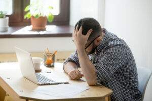 Man sitting at desk in front of laptop with head in hands, tired and frustrated